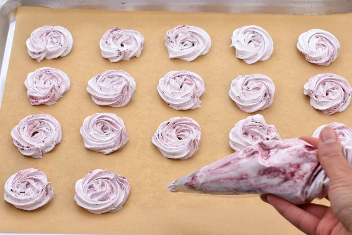 hand holding piping bag with meringue cookies on baking sheet.