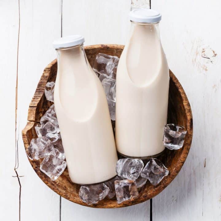 two glass containers of whole milk sitting in ice in a wooden bowl.
