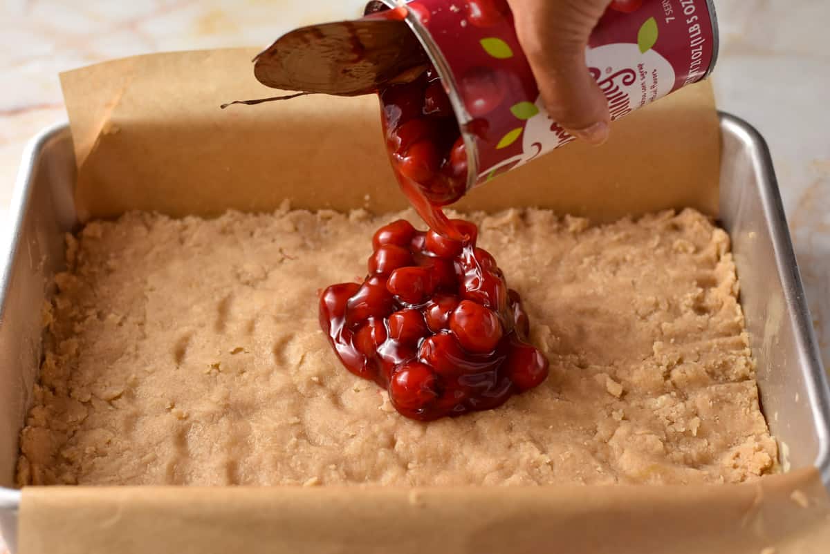 hand pouring cherry pie filling onto crust.