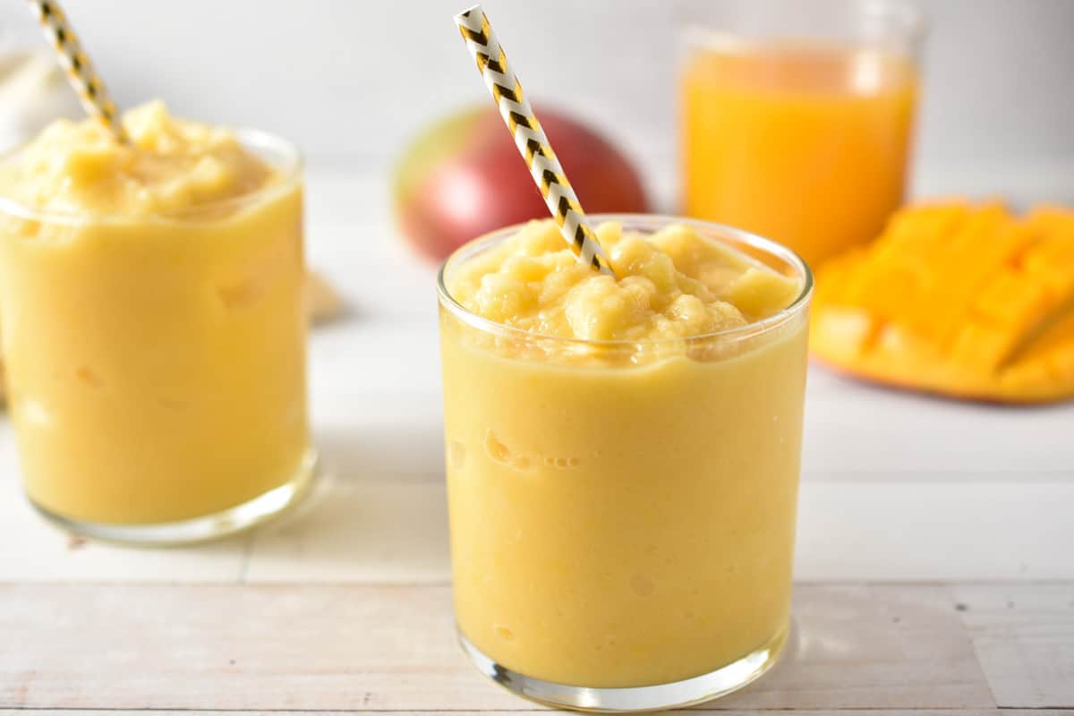 two mango smoothie glasses with a straw, with diced mango and a glass of juice in the background. 