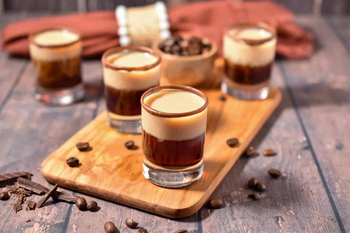 four shot glasses with layers of liquor on a wooden cutting board with coffee beans. 
