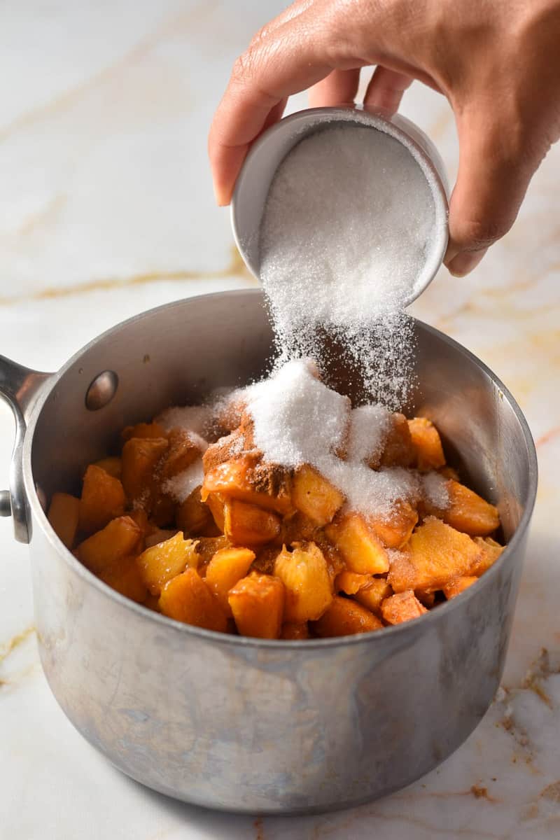 hand pouring sugar into a pan of diced peaches. 