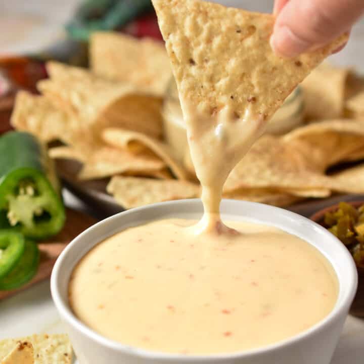 A small bowl with queso served on a plate with tortilla chips. A tortilla chip is dipped in the queso.