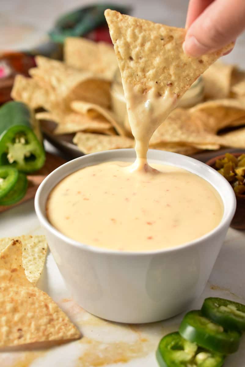 queso in a bowl with jalapenos and tortilla chips. a hand is dipping a chip into the bowl. 