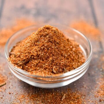 A small glass bowl filled with a reddish-brown Hello Fresh Tunisian Spice Blend sits on a wooden surface. Some of the aromatic spice mix spills outside the bowl, revealing its coarse texture and visible herbs.