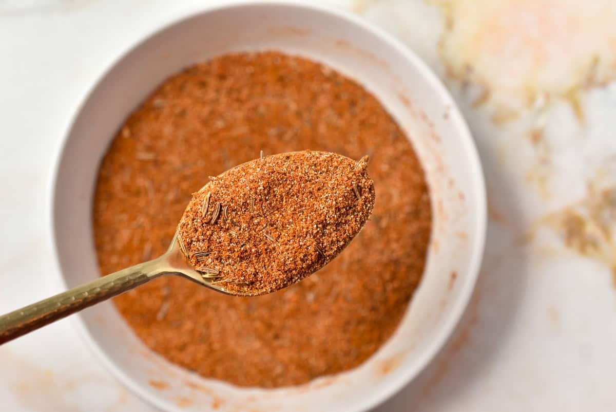 A spoonful of Hello Fresh Tunisian Spice Blend hovers above a white bowl brimming with the aromatic, reddish-brown mix, set against a light, marble-like surface.