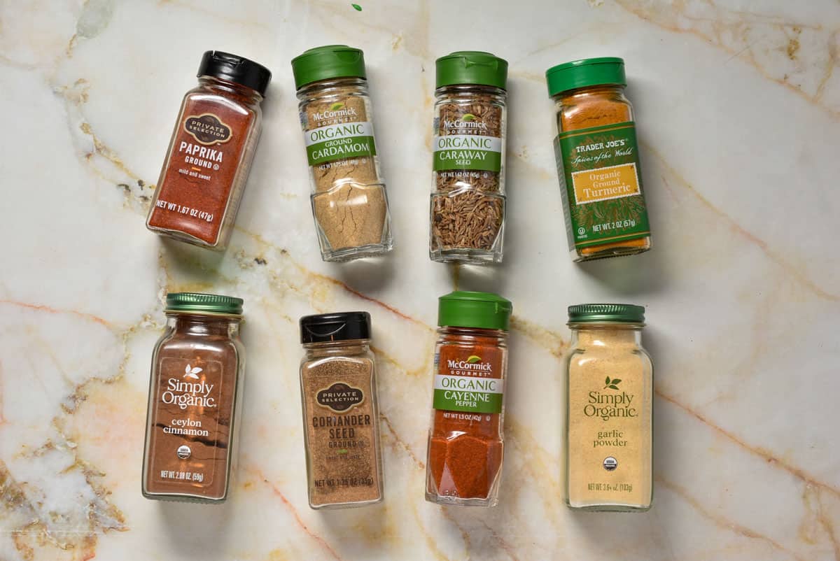 A collection of spice jars, used to make the flavorful Hello Fresh Tunisian Spice Blend, arranged on a marble surface. Featuring paprika, cardamom, and more, each jar is topped with a green or brown cap and labeled clearly for your culinary adventures.