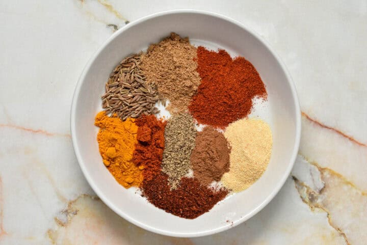 A white plate showcases an array of spices-cumin seeds, ground cinnamon, chili powder, paprika, turmeric, black pepper, garlic powder, and ground coriander-each artfully arranged in separate piles on a light marbled background reminiscent of a Hello Fresh Tunisian Spice Blend.