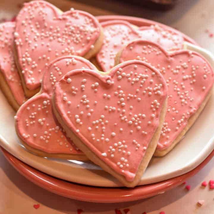 heart shaped cookie with pink royal icing and white sprinkles sitting on a metal baking sheet.