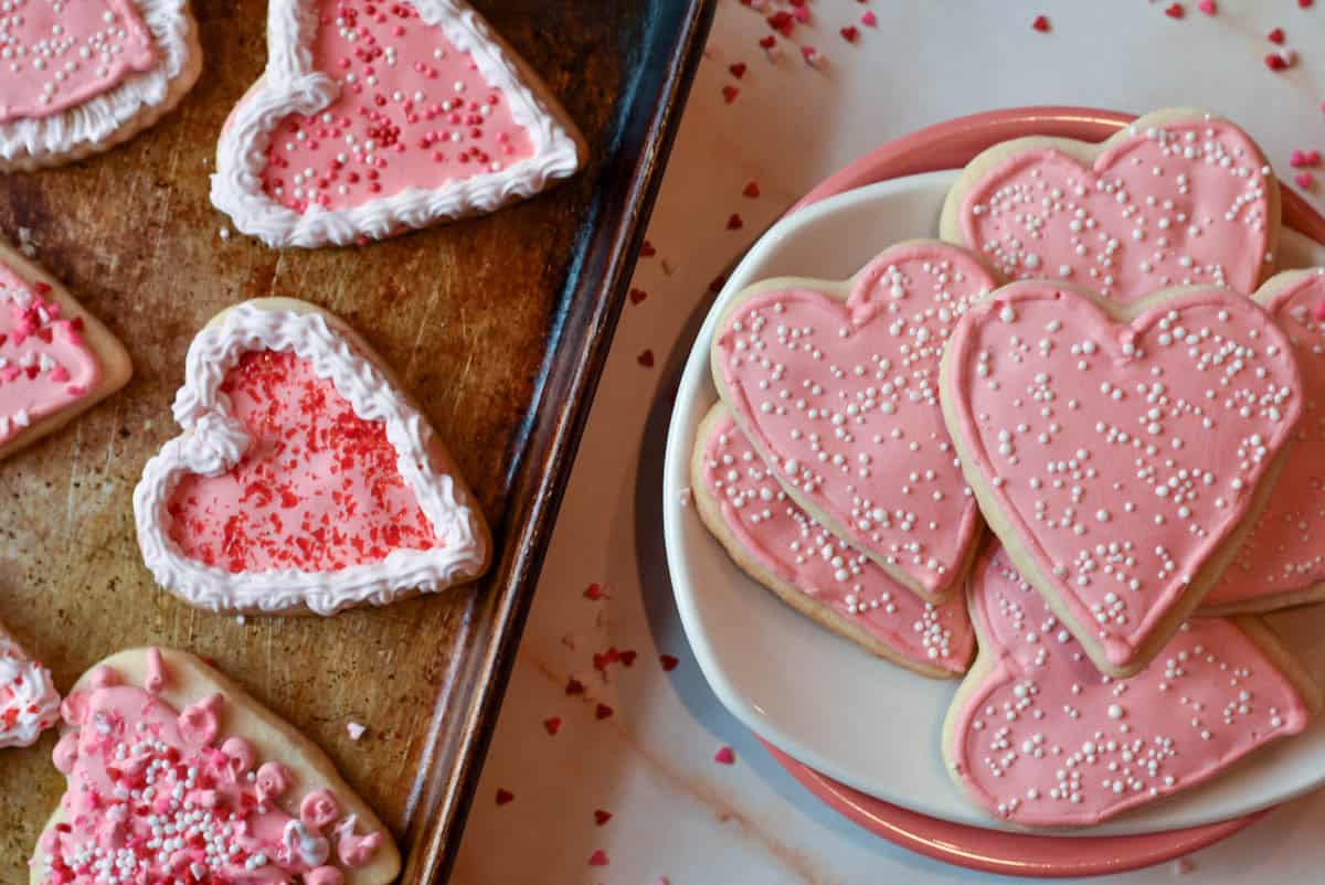 A plate of heart-shaped sugar cookies with royal icing in soft pastel hues and sprinkled with festive decorations.