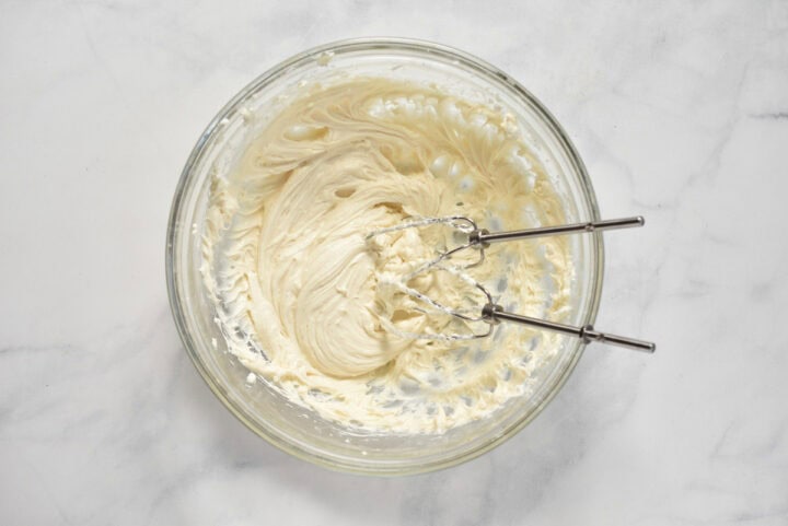 A glass bowl filled with creamy, whipped frosting and two mixer beaters resting inside on a marble surface, reminiscent of the smooth topping on a cherry danish.