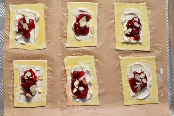 Six rectangular cherry danishes with cream cheese filling and almond slices rest on parchment paper, ready for baking.