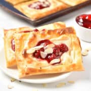 A pair of cherry danishes, elegantly adorned with almond slices, rest on a white plate. In the background, a tempting array of pastries and vibrant cherry filling awaits discovery.