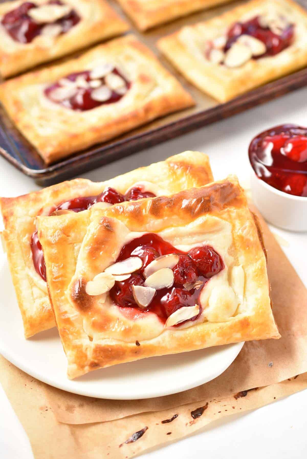 Cherry danishes with almond slices rest elegantly on a plate beside a bowl of rich cherry filling. In the background, additional pastries await on a baking sheet, promising sweet indulgence.