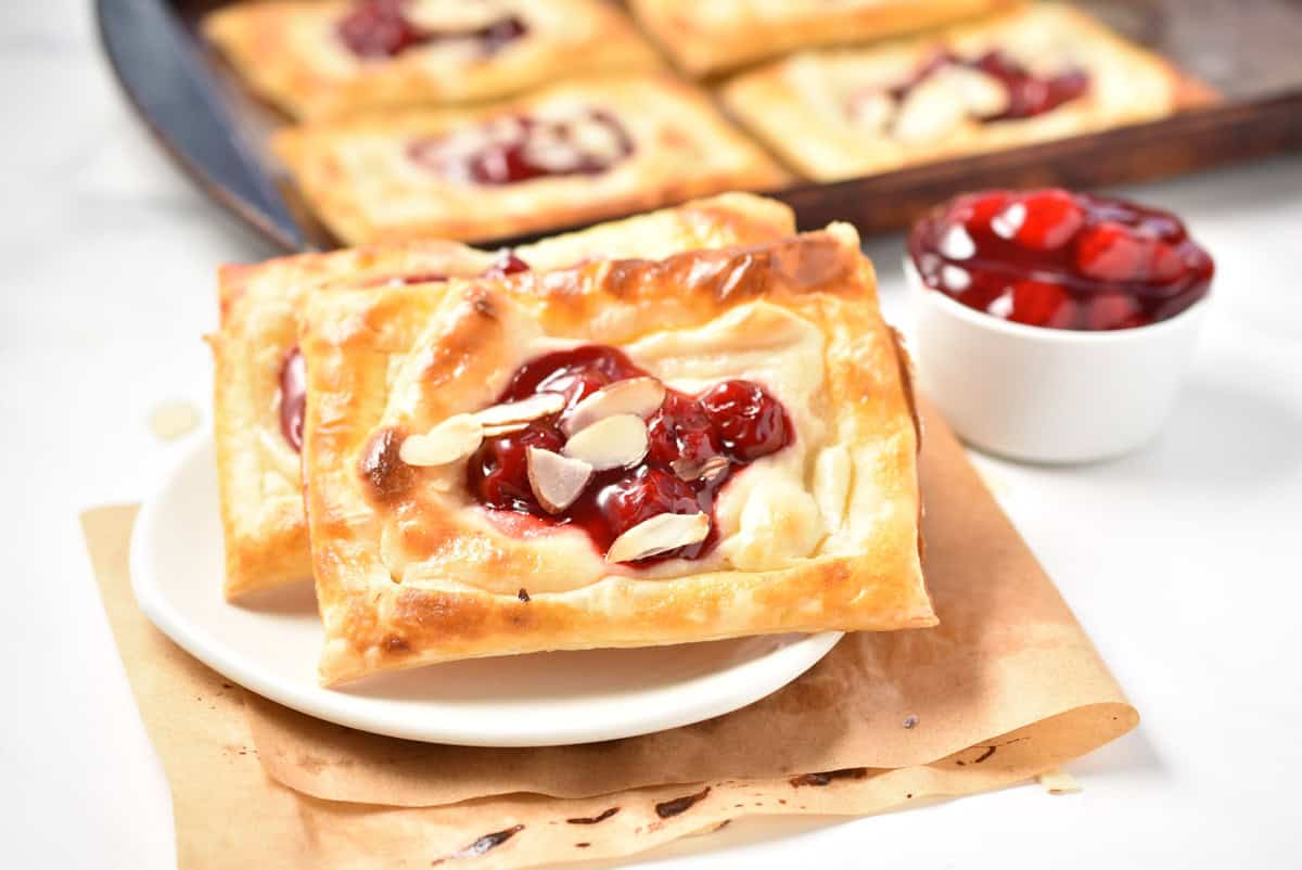 A cherry Danish is placed artfully on a plate, its pastry adorned with almond slices and a luscious cherry filling, accompanied by a small bowl of cherries.
