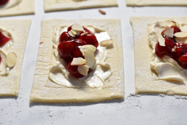 A cherry Danish masterpiece: rectangular pastry dough, elegantly spread with cream cheese and rich cherry filling, crowned with delicate almond slices on a floured surface.