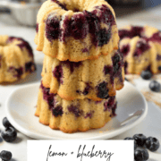 A stack of delightful lemon blueberry mini bundt cakes sits on a white plate, adorned with fresh blueberries. In the softly blurred background, a baking tray showcases even more cakes. The text reads Lemon + Blueberry Mini Bundt Cakes alongside a handy website link.