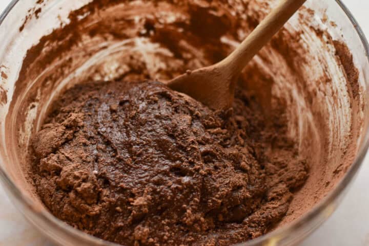 A brown, lumpy batter mixture for air fryer brownies is being stirred with a wooden spoon in a clear glass bowl.