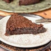 A brownie slice rests on a white plate dusted with cocoa powder. A fork awaits beside it. In the background, a large platter offers more tempting slices, each promising delightful indulgence.