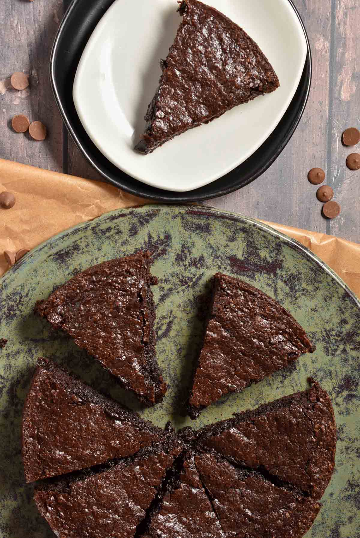 A chocolate brownie cake, fresh from the air fryer, is cut into five slices on a round greenish plate. One slice rests on a white plate with a black rim above. The background is adorned with scattered chocolate chips and a brown paper sheet.