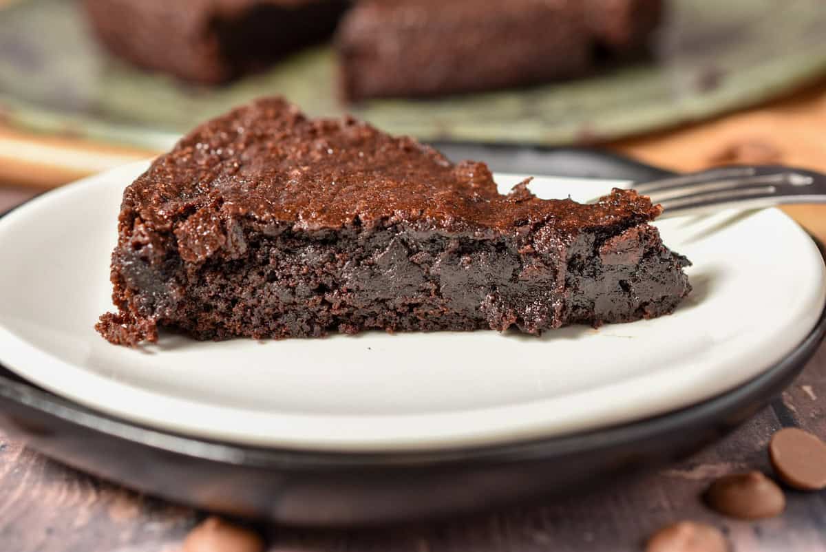A brownie slice, sits on a white plate with a dark rim. Its dense texture and deep brown hue invite indulgence. A fork peeks from the side.