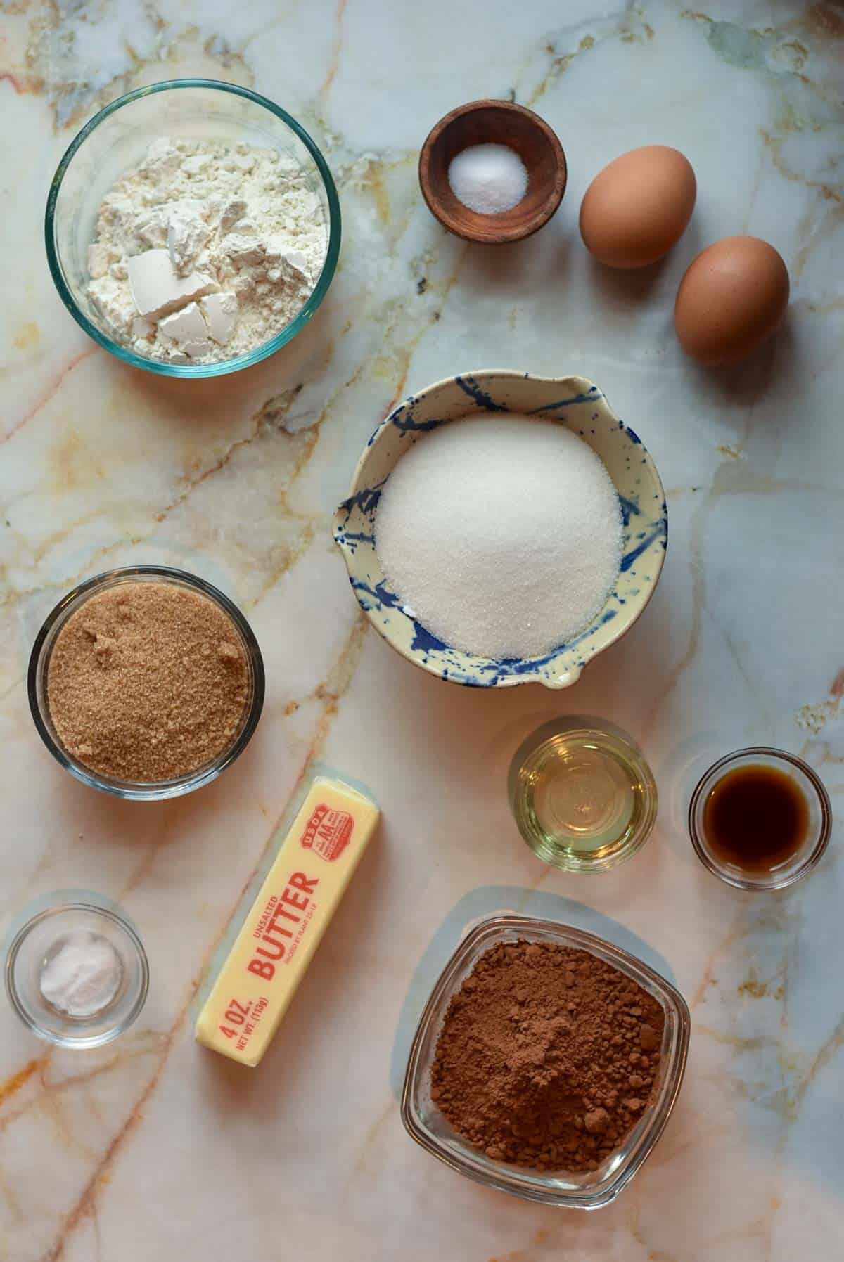 A marble countertop showcases the essential baking ingredients for brownies: flour in a glass bowl, two eggs, sugar, brown sugar, a stick of butter, cocoa powder, vegetable oil in a small bowl, vanilla extract, salt, and baking soda in small containers.