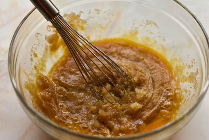 A whisk rests in a glass bowl containing a mixture of brown sugar and melted butter.