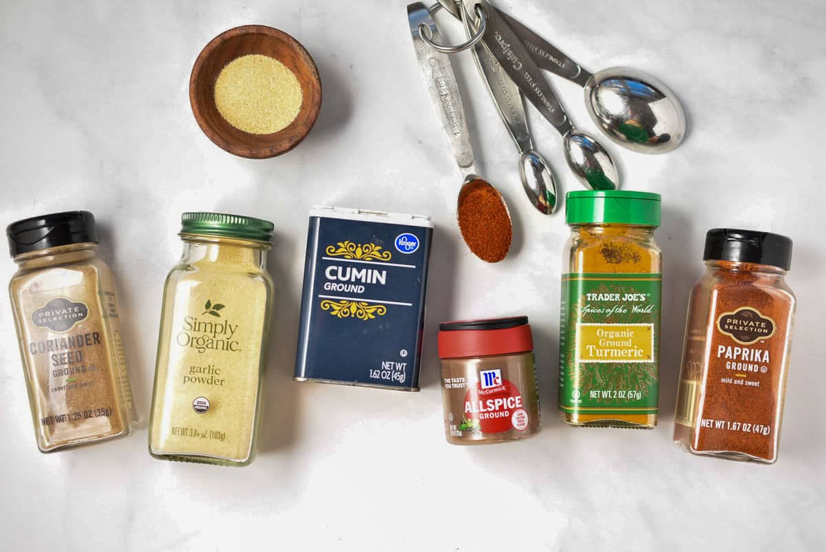 A vibrant display of various spice containers and measuring spoons on a white surface, including coriander, garlic powder, cumin, allspice, turmeric, and paprika-a perfect setup for creating your own shawarma spice blend.