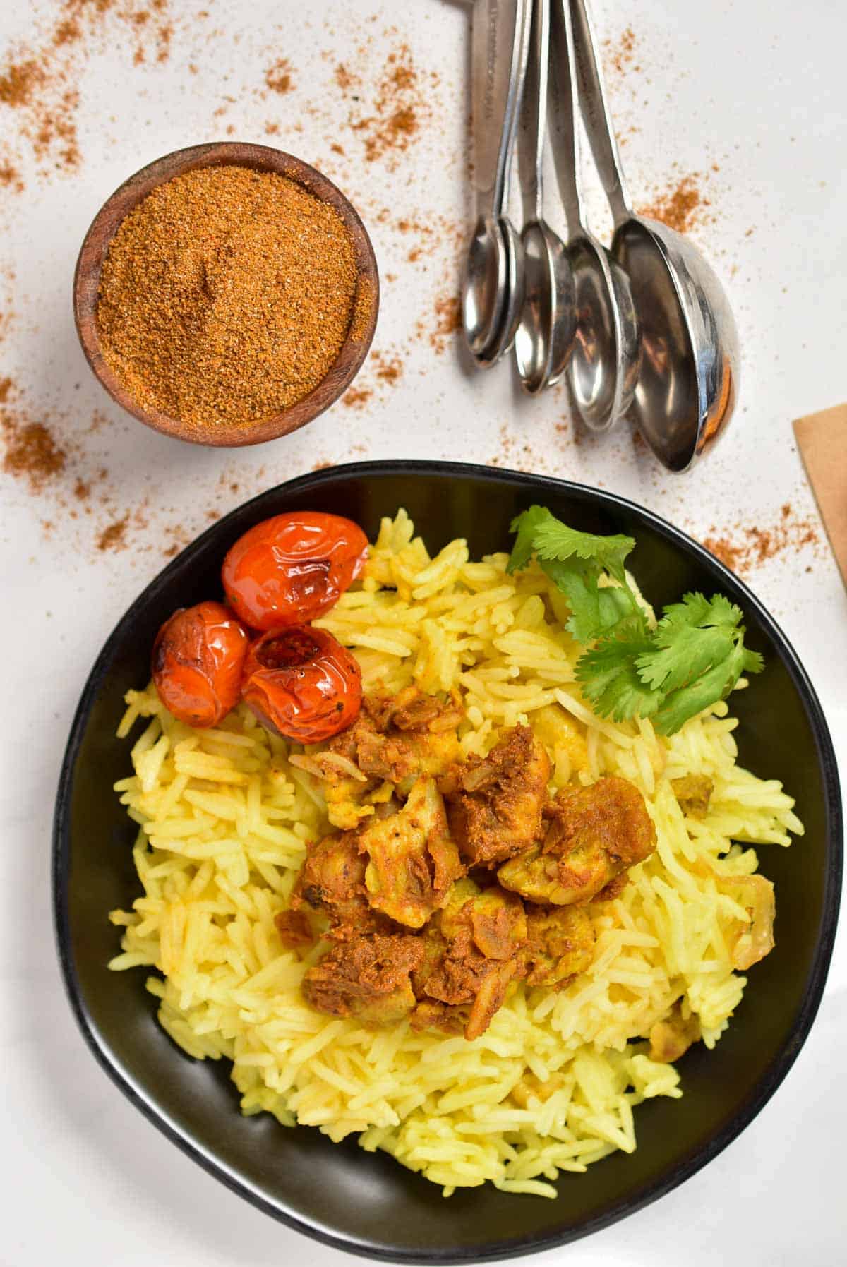 A black bowl features yellow rice, spiced meat, cherry tomatoes, and cilantro, showcasing the flavors of a Shawarma Spice Blend. Nearby, a wooden bowl of spice and Hello Fresh measuring spoons rest on a white surface, ready to enhance your culinary creations.