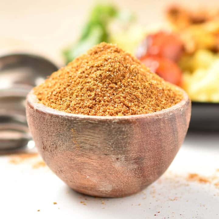 A wooden bowl brimming with shawarma spice takes center stage on a white surface, surrounded by scattered spices. In the blurred background, metal spoons and food hint at a Hello Fresh culinary journey waiting to unfold.