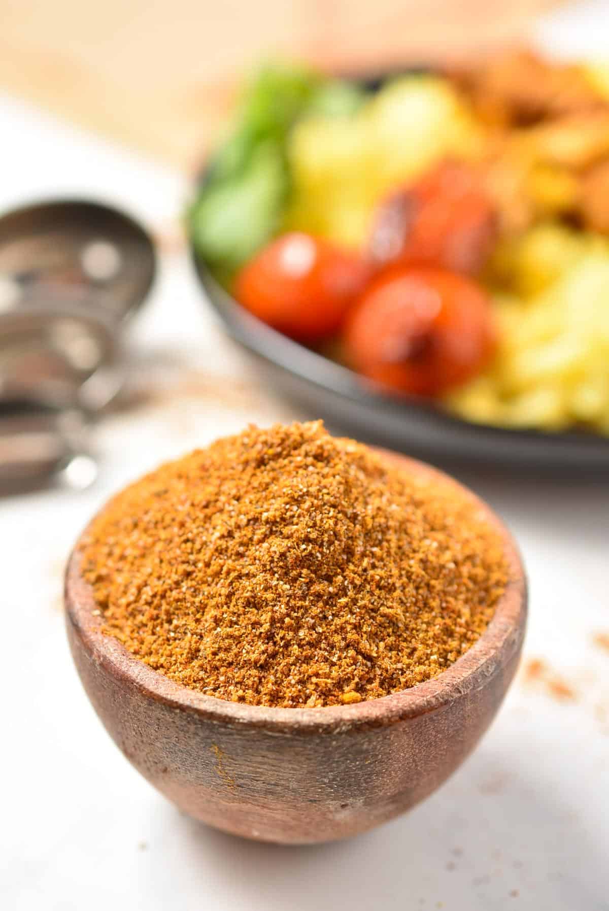 A wooden bowl filled with a delectable Shawarma spice blend is in the foreground. In the background, a plate with tomatoes and greens is slightly blurred.