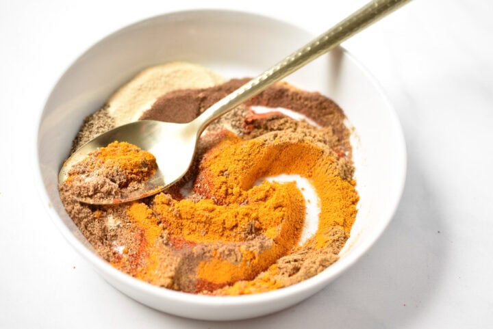 A white bowl contains a Shawarma Spice Blend of orange, brown, and beige spices, with a gold spoon resting inside.