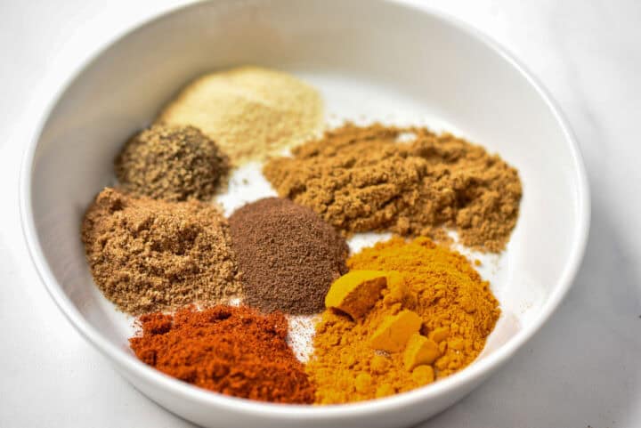A white bowl holds a rainbow of seven ground spices in small piles, each boasting unique colors and textures.