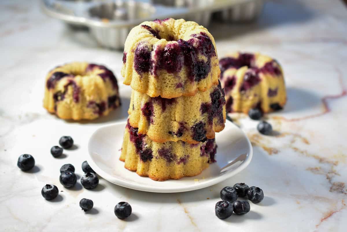 Three mini bundt cakes, infused with zesty lemon blueberry flavors, are stacked gracefully on a white plate. Additional blueberries are sprinkled artfully around the marble surface, while another bundt pan sets the scene in the background.