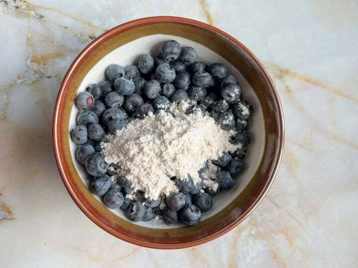 A bowl filled with fresh blueberries and topped with flour. The bowl has a light brown rim and beige interior, perfectly complementing the vibrant blueberries.