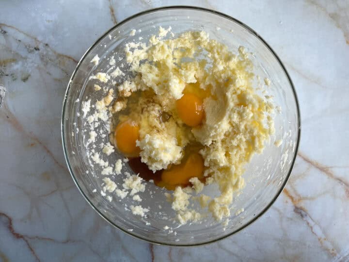A glass bowl on a marble surface contains a mix of creamed butter and sugar with three cracked eggs nestled on top, ready to be mixed further.