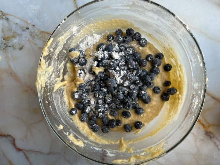 A glass bowl on a marble countertop contains lemon blueberry muffin batter. Fresh blueberries, lightly dusted with flour, are mixed in. The batter has a creamy texture with some visible flour on the fruit, ready to transform into delightful mini bundt cakes.