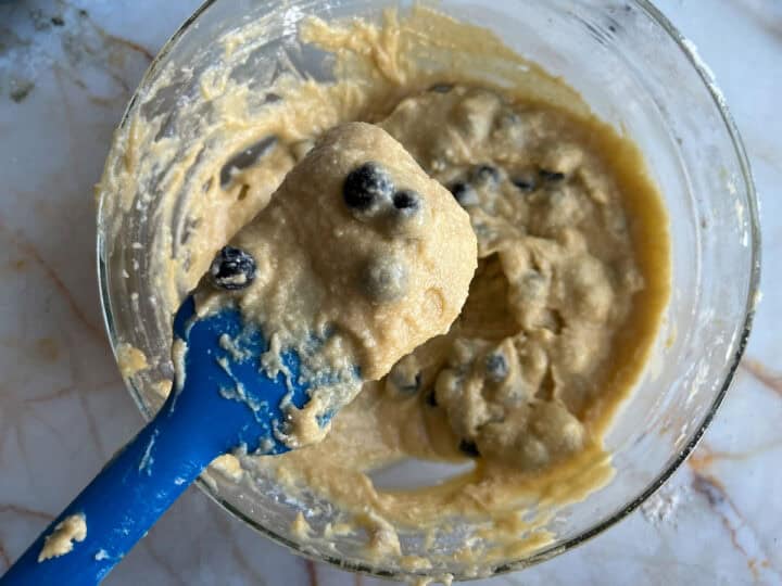 A glass bowl with batter rests on a marble surface. The batter, rich with blueberries and a hint of zesty lemon, is being mixed with a blue spatula that lifts a portion of the delightful mixture.