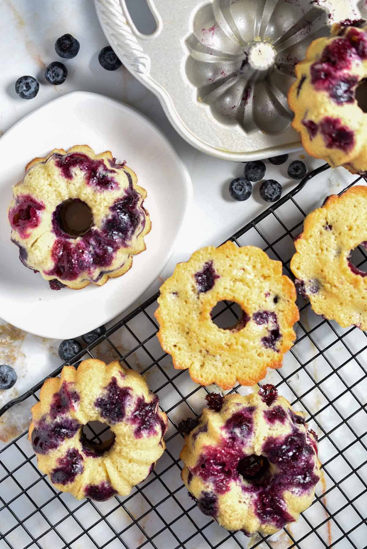 Lemon blueberry mini bundt cakes with vibrant berries are displayed on a cooling rack and a white plate. A baking mold dusted with batter traces and scattered blueberries completes the delectable scene on the marble surface.