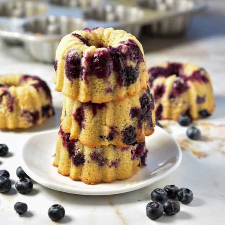 Three mini blueberry bundt cakes are stacked on a white plate, their tops glistening with a hint of lemon. Fresh blueberries are scattered across the marble surface, adding a pop of color, while a baking tray lingers in the background.