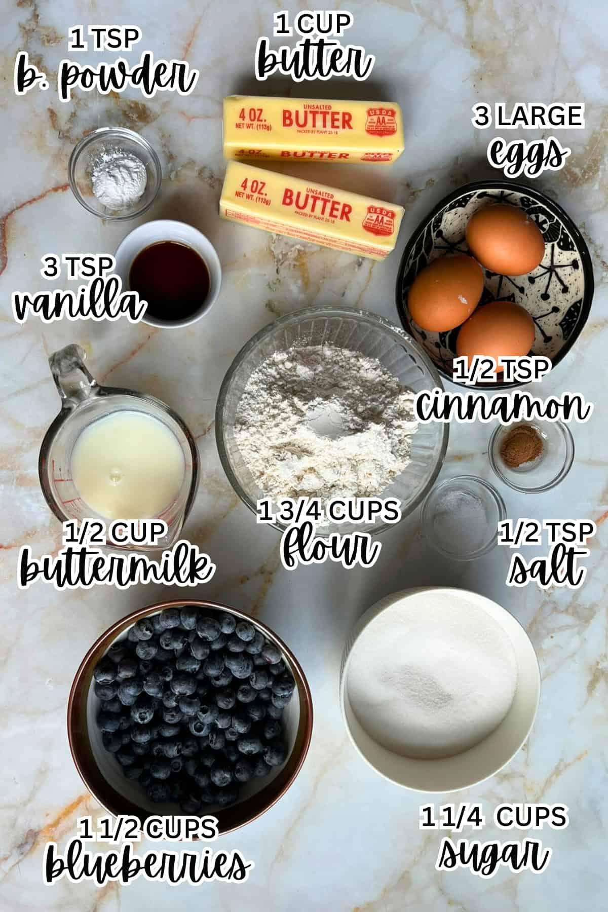 Top-down view of baking ingredients on a marble surface for a Lemon Blueberry Mini Bundt Cake: butter, eggs, blueberries, flour, sugar, buttermilk, baking powder, vanilla, cinnamon, lemon zest, and salt. Each item is labeled with its quantity.