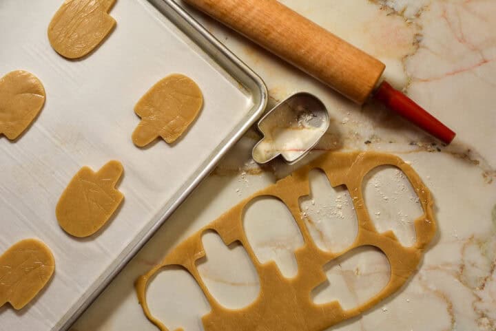 A baking sheet with mitten-shaped sugar cookies cutouts, a rolling pin, a mitten-shaped cookie cutter, and rolled dough on a marble surface-perfect for festive holiday desserts.