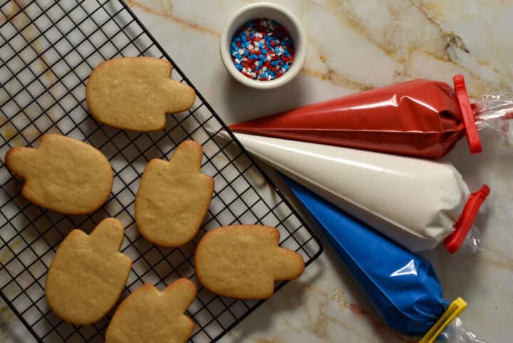Unfrosted mitten-shaped sugar cookies on a cooling rack sit next to piping bags of red, white, and blue icing and a bowl of star-shaped sprinkles-perfect for making festive Fourth of July cookies.