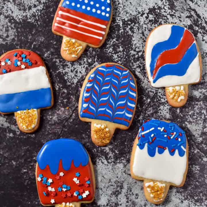 Six popsicle-shaped fourth of July sugar cookies are decorated with red, white, and blue icing and sprinkles, arranged on a dark textured surface.