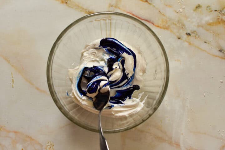 A glass bowl with white frosting and blue food coloring being mixed with a spoon on a marble surface, perfect for decorating festive Fourth of July cookies.
