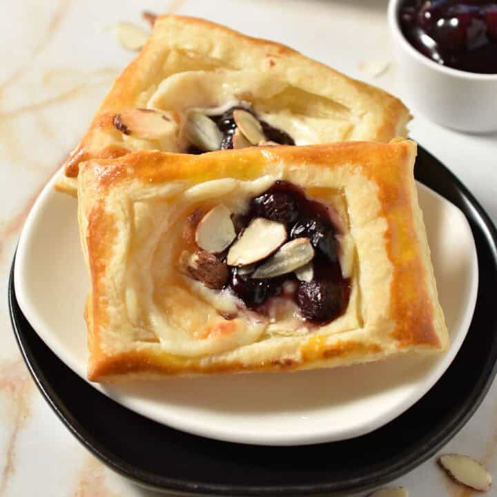 Two rectangular pastries, reminiscent of a Blueberry Danish, with cheese and cherry filling and sliced almonds, are served on a white plate. A small bowl of cherry filling is in the background.