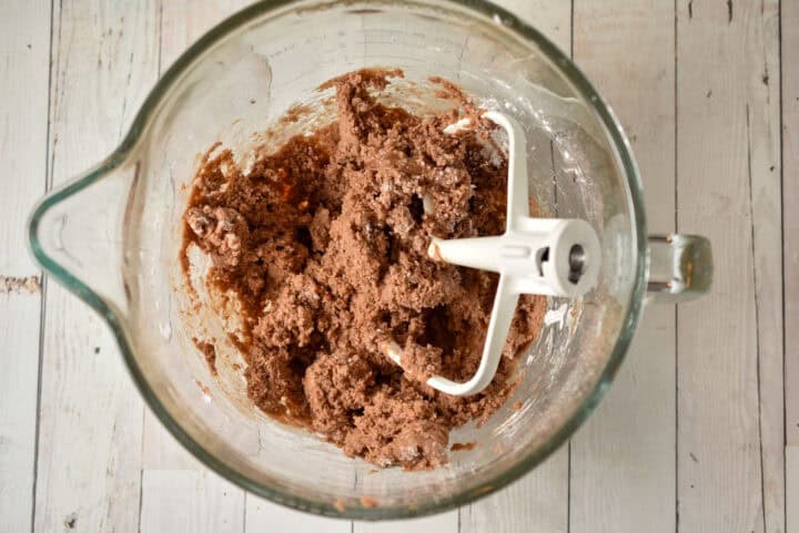 A glass mixing bowl containing partially mixed Potato Fudge cookie dough with a white paddle attachment, seen from above on a light wooden surface.
