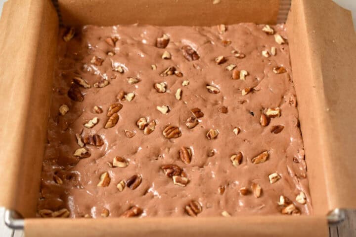 A square baking pan lined with parchment paper is filled with chocolate batter, inspired by a classic potato fudge recipe, and topped with chopped pecans, ready to be baked.