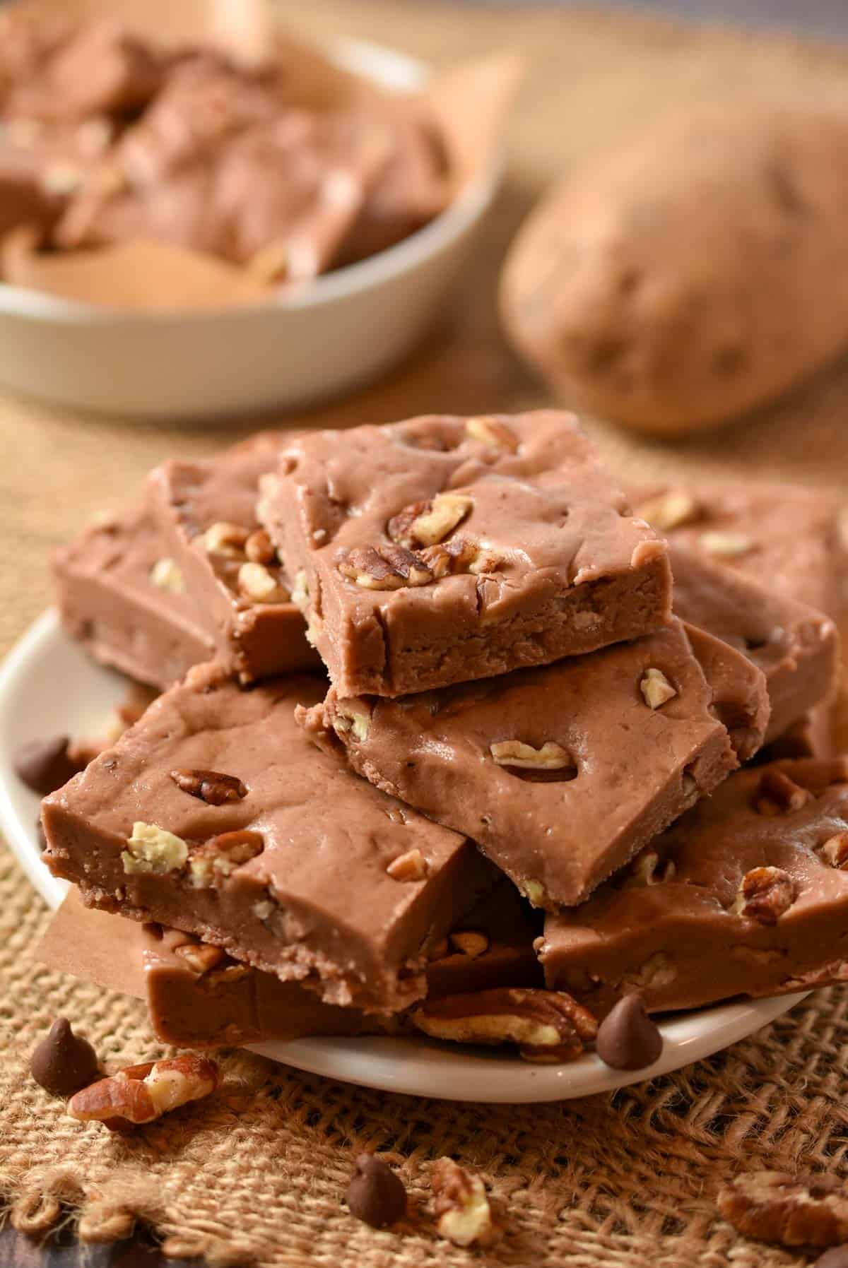 A stack of chocolate fudge squares with visible nut pieces sits on a white plate, showcasing this unique Potato Fudge recipe, with more fudge in a bowl and a potato in the background.