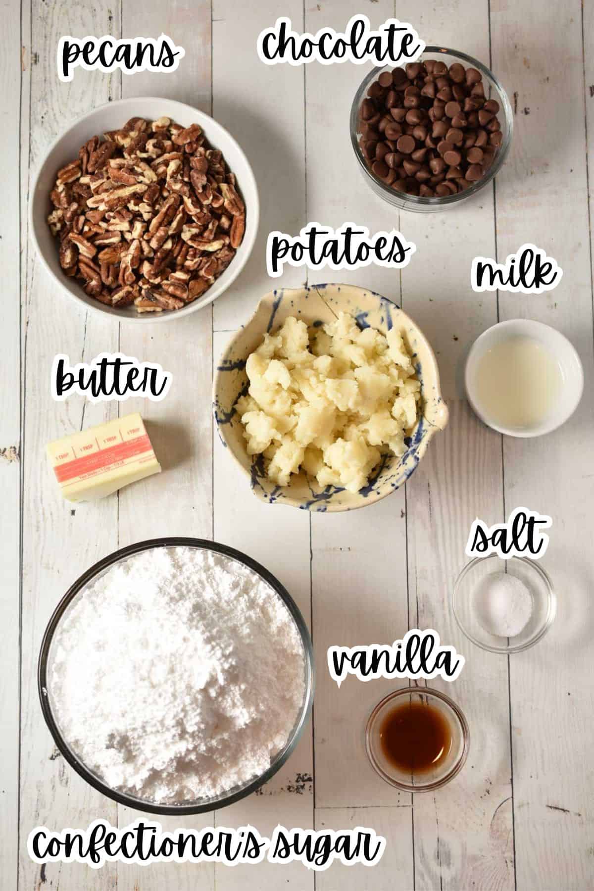 Bowls on a white surface containing ingredients for Potato Fudge-pecans, chocolate chips, mashed potatoes, milk, butter, salt, vanilla extract, and confectioners sugar-each labeled with text for this classic fudge recipe.
